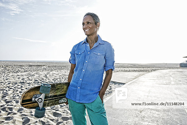 Ältere schwarze Frau hält Skateboard am Strand