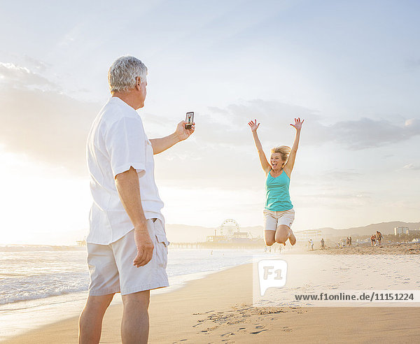 Kaukasischer Mann fotografiert seine Frau am Strand mit dem Handy