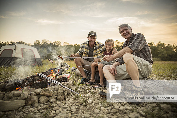 Three generations of Caucasian men roasting hot dogs over campfire