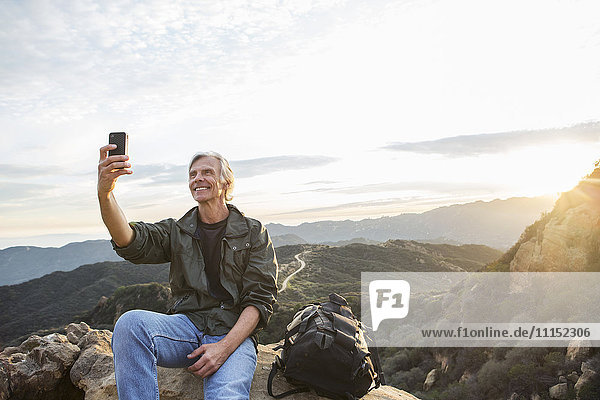 Older Caucasian man taking cell phone photograph on rocky hilltop