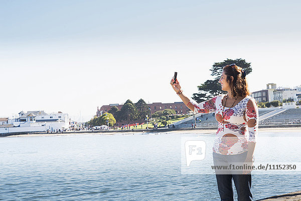Hispanische Frau macht Selfie mit ihrem Handy am Wasser