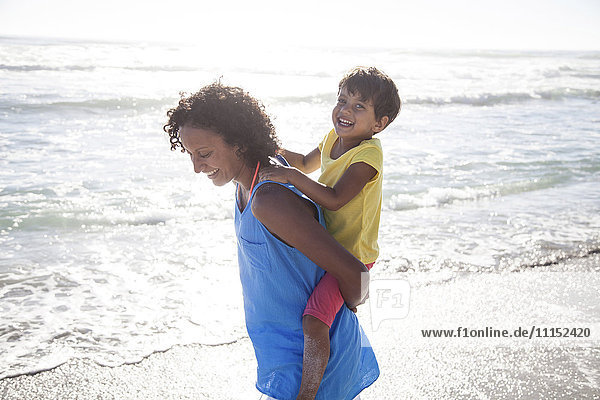Gemischtrassige Mutter trägt Tochter huckepack am Strand