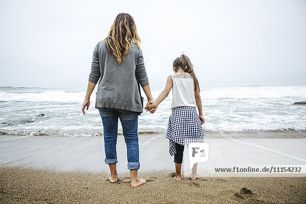 Hispanic mother and daughter standing barefoot on beach