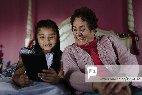 Hispanische Großmutter und Enkelin benutzen digitales Tablet auf dem Bett