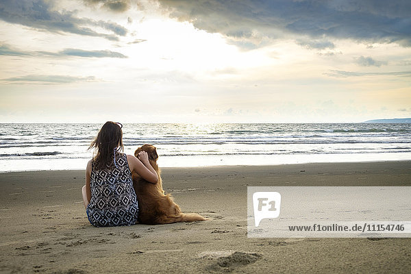 Mexiko  Nayarit  Rückansicht einer jungen Frau  die neben ihrem Hund am Strand sitzt.