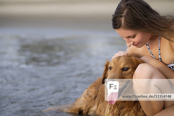 Nahaufnahme einer jungen Frau im Bikini  die ihren Golden Retriever Hund am Strand streichelt.