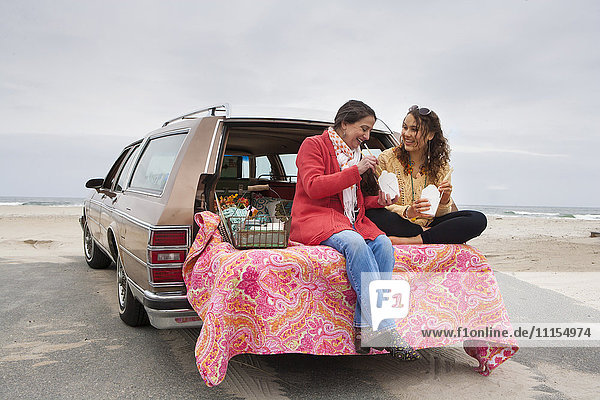 Kaukasische Mutter und Tochter beim Picknick im Kofferraum eines Autos am Strand