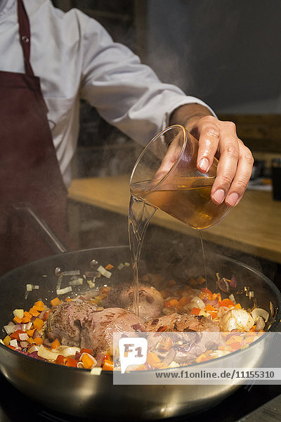 Man pouring a glass of brandy on beef cheeks in a pan with sauteed vegetables