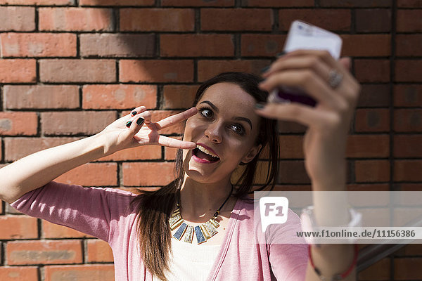 Portrait of young woman showing victory sign taking selfie with smartphone