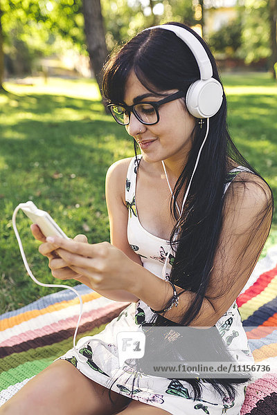 Junge Frau sitzt auf einer Decke im Park und hört Musik.