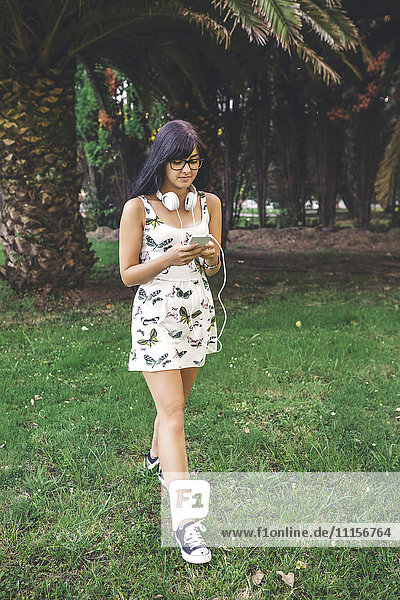 Junge Frau mit Kopfhörern  die im Park spazieren geht und auf das Handy schaut.