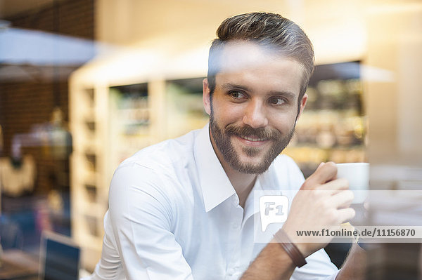 Lächelnder Geschäftsmann hält eine Tasse Kaffee hinter einer Fensterscheibe in einem Café.