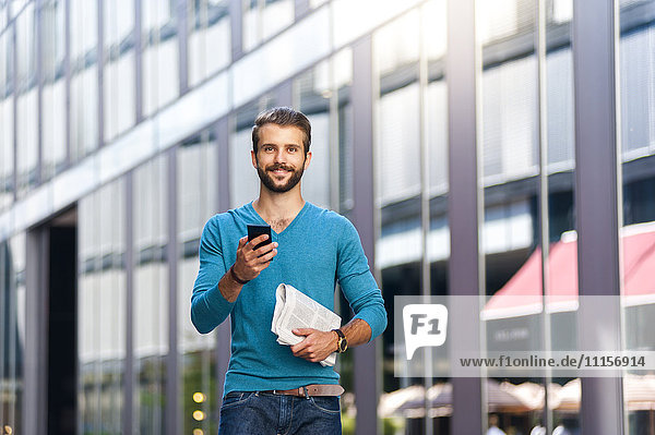 Lächelnder junger Mann mit Handy und Zeitung in der Stadt