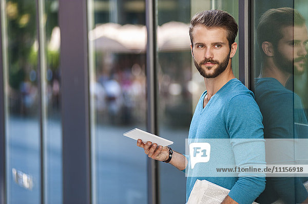 Junger Mann mit Tablette und Zeitung an der Glasfront eines Gebäudes