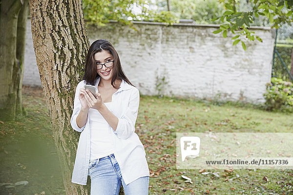 Junge Frau mit Smartphone an Baumstamm gelehnt