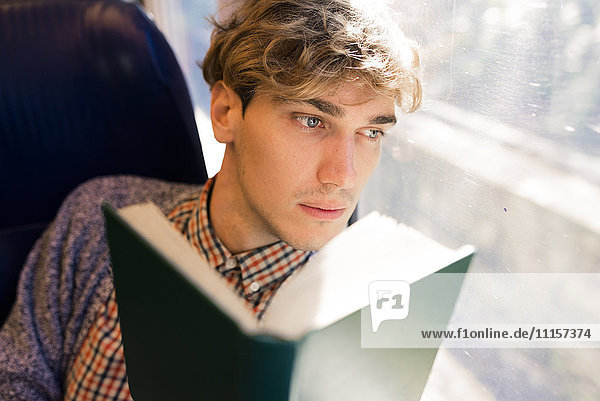 Nachdenklicher junger Mann mit Buch im Zug und Blick durchs Fenster