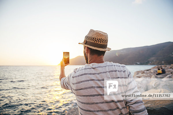 Griechenland  Cylcades Inseln  Amorgos  Mann fotografiert den Sonnenuntergang mit einem Smartphone am Meer.
