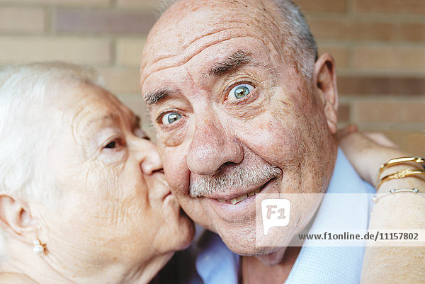 Ein älterer Mann zieht ein komisches Gesicht  während seine Frau ihn küsst.