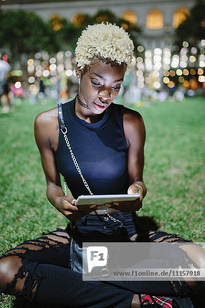 Young woman sitting on meadow of a park at night using tablet