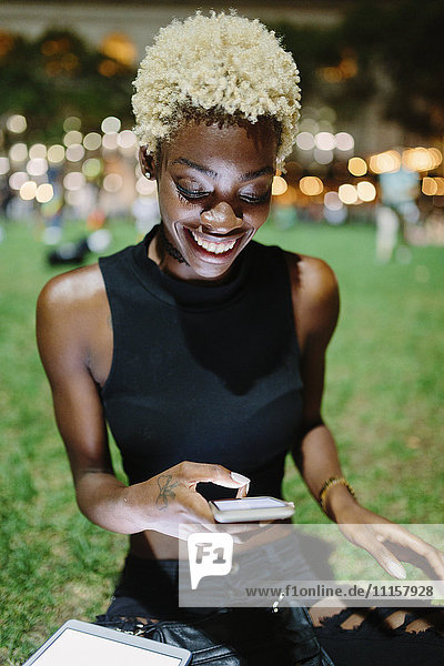 Smiling young woman sitting on meadow of a park at night text messaging
