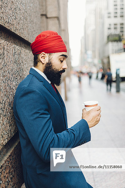 Indischer Geschäftsmann in Manhattan lehnt sich an die Wand und trinkt Kaffee.