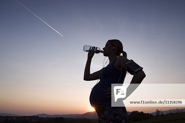 Silhouette einer schwangeren Frau  die bei Sonnenuntergang Wasser aus der Flasche trinkt.