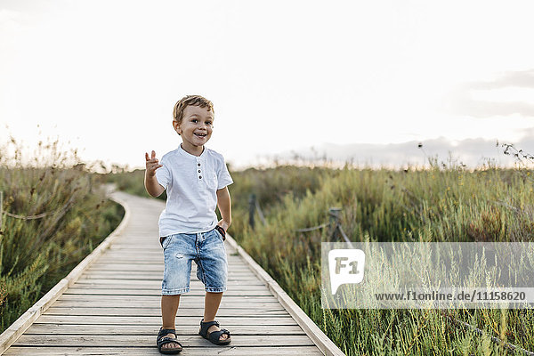 Fröhlicher kleiner Junge steht auf einer Strandpromenade in der Natur.