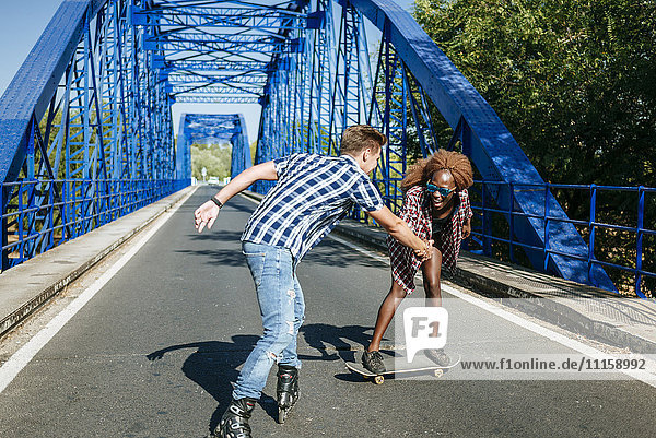 Glückliches junges Paar mit Inline-Skates und Skateboard auf einer Brücke
