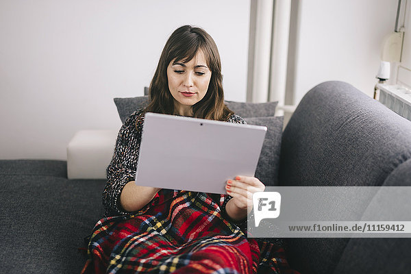 Junge Frau mit einer Decke bedeckt  die sich mit ihrer Tablette auf der Couch entspannt.
