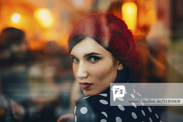 Porträt einer jungen Frau mit rotem Hut,  die abends durch das Fenster eines Pubs schaut.