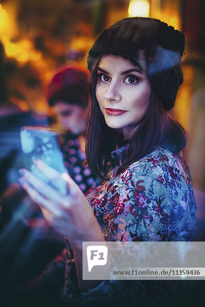 Porträt einer jungen Frau mit Smartphone beim Blick durch die Fensterscheibe einer Kneipe am Abend