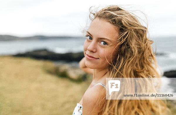 Porträt eines lächelnden blonden Mädchens mit blasenden Haaren an der Küste