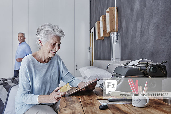 Lächelnde Seniorin online einkaufen im Schlafzimmer mit Ehemann im Hintergrund