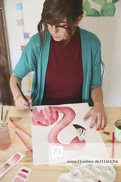 Frau malt Aquarell eines Flamingos auf dem Schreibtisch in ihrem Atelier