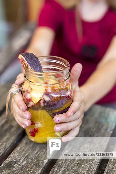 Hände eines Mädchens  das ein Glas Wasser mit verschiedenen Früchten hält.