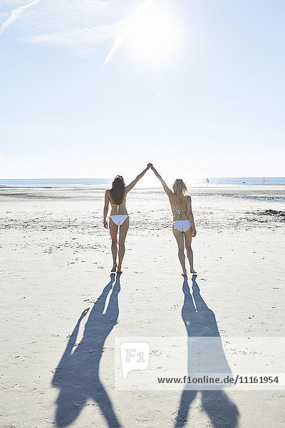 Zwei Freundinnen im Bikini gehen Hand in Hand am Strand.