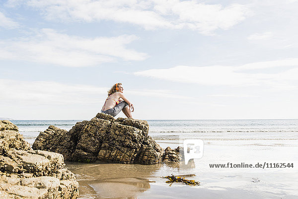 Teenager-Mädchen sitzt auf einem Felsen am Strand und hört Musik.