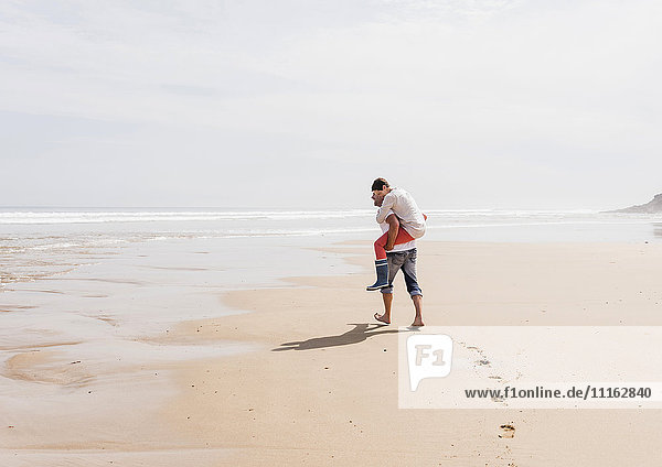Erwachsener Mann  der seine Frau huckepack am Strand trägt.