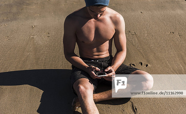 Junger Mann mit Handy am Strand sitzend