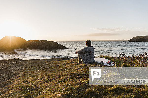 Frankreich  Bretagne  Halbinsel Crozon  Frau bei Sonnenuntergang an der Küste sitzend mit Surfbrett