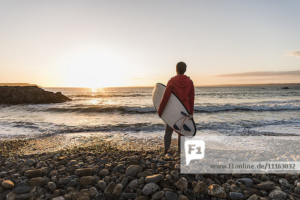 Frankreich  Bretagne  Halbinsel Crozon  Frau steht am steinigen Strand bei Sonnenuntergang mit Surfbrett