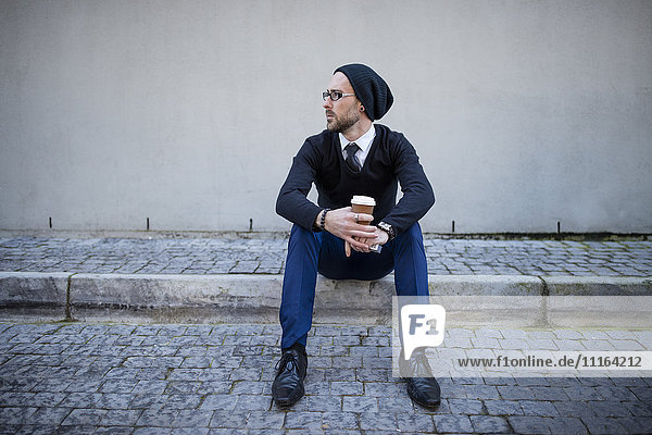 Junger Mann auf dem Bordstein sitzend mit Kaffee zum Mitnehmen