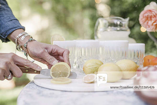 Nahaufnahme einer Frau,  die an einem Tisch in einem Garten steht und für ein Getränk Zitronen in Scheiben schneidet.