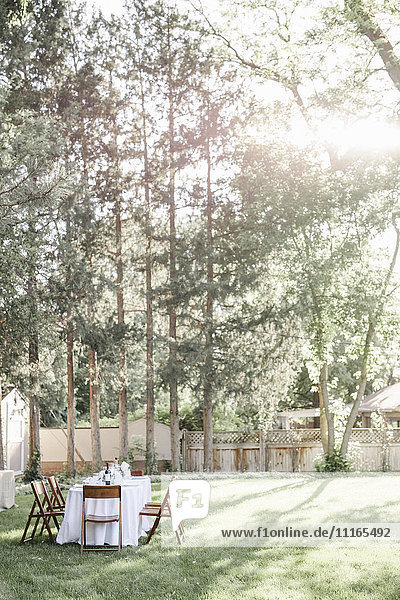 Gedeckter Tisch mit einer weißen Tischdecke und Holzstühlen in einem Garten.