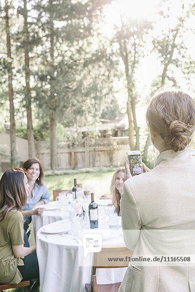 Eine Frau fotografiert mit ihrem Mobiltelefon eine Gruppe von Frauen  die an einem Tisch in einem Garten sitzen.