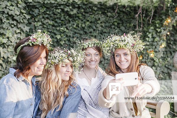 Vier lächelnde Frauen mit Blumenkränzen im Haar  die in einem Garten sitzen und sich ein Selfie nehmen.