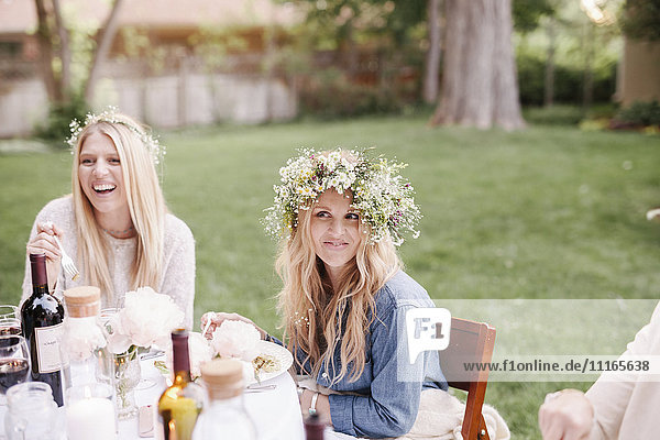 Zwei lächelnde Frauen mit Blumenkränzen im Haar  die an einem Tisch in einem Garten sitzen.