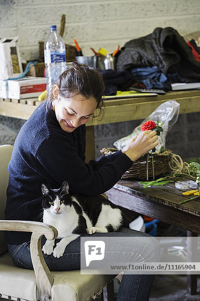 Eine Frau in einer Blumenscheune  die mit einer auf ihrem Schoß sitzenden Katze an einem Arrangement arbeitet.