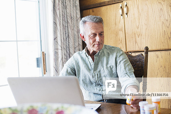 Älterer Mann sitzt an einem Tisch  benutzt einen Laptop-Computer und hält eine Flasche Pillen in der Hand.