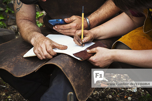 Man and woman  blacksmiths wearing aprons writing into a notebook sat in a garden.
